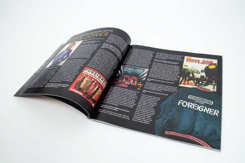 An open tour book spread with pages that are mostly black with several printed photos, text blocks, and colorful album-cover style graphics, including the word FOREIGNER prominently visible on the right page.